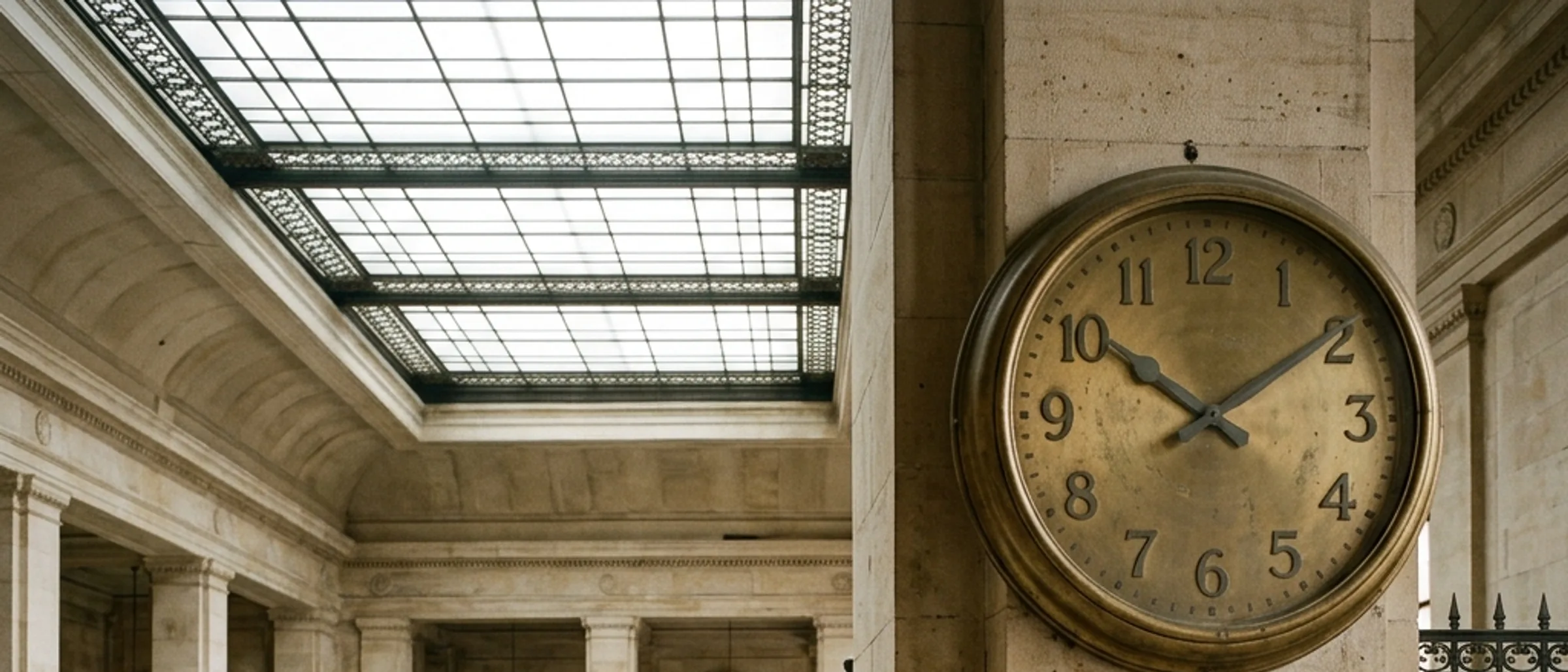 An old brass wall clock inside a grand bank hall with stone pillars.